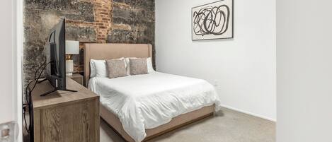 Un dormitorio con cama, televisor sobre un mueble de madera, pared de ladrillo y una obra de arte abstracta.