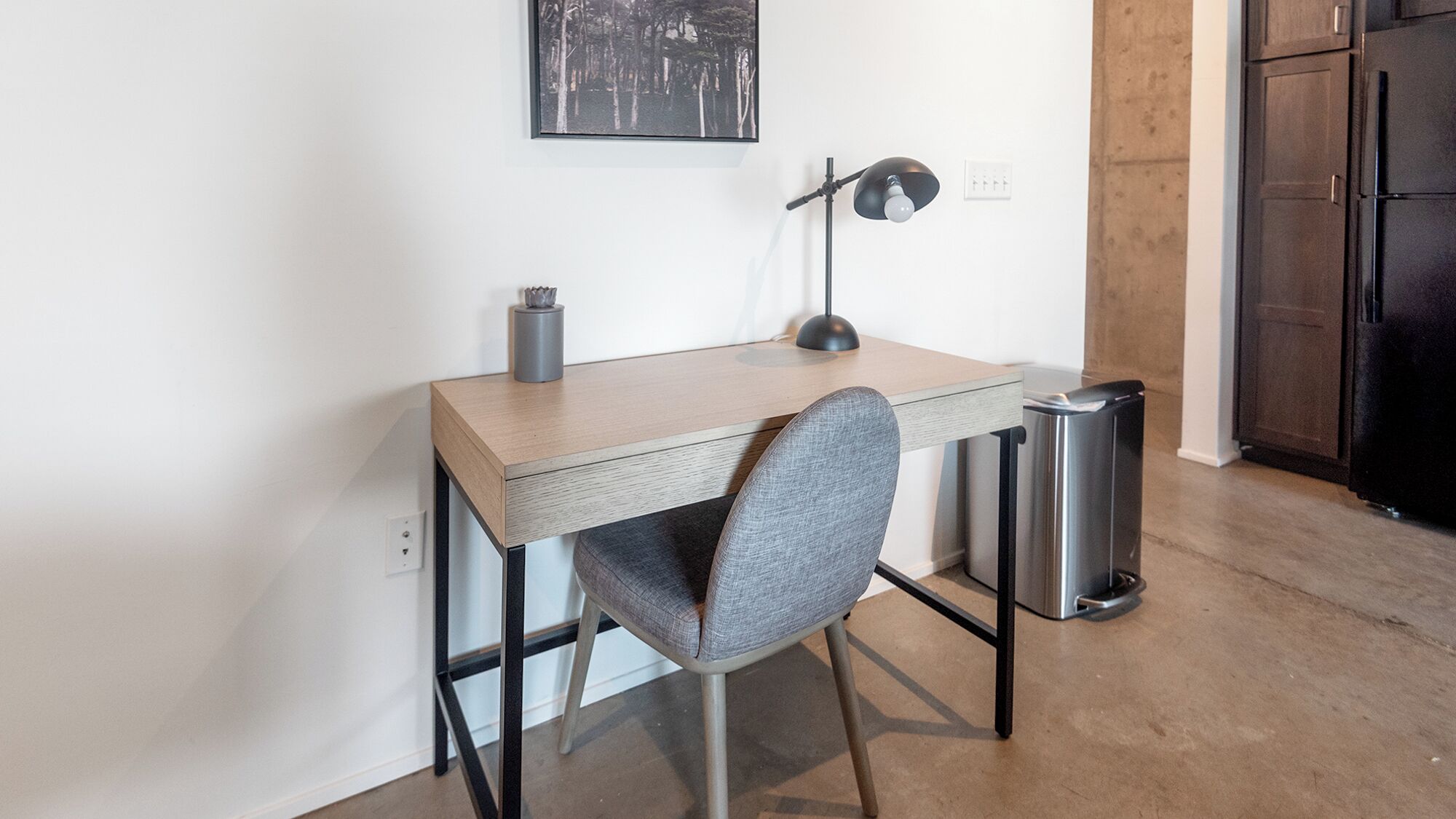 Een compacte, moderne werkplek met een houten bureau, een grijze stoel, een bureaulamp en een prullenbak.