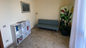 Ett rum med en blå soffa, en vit bokhylla, en tavla och en stor krukväxt.