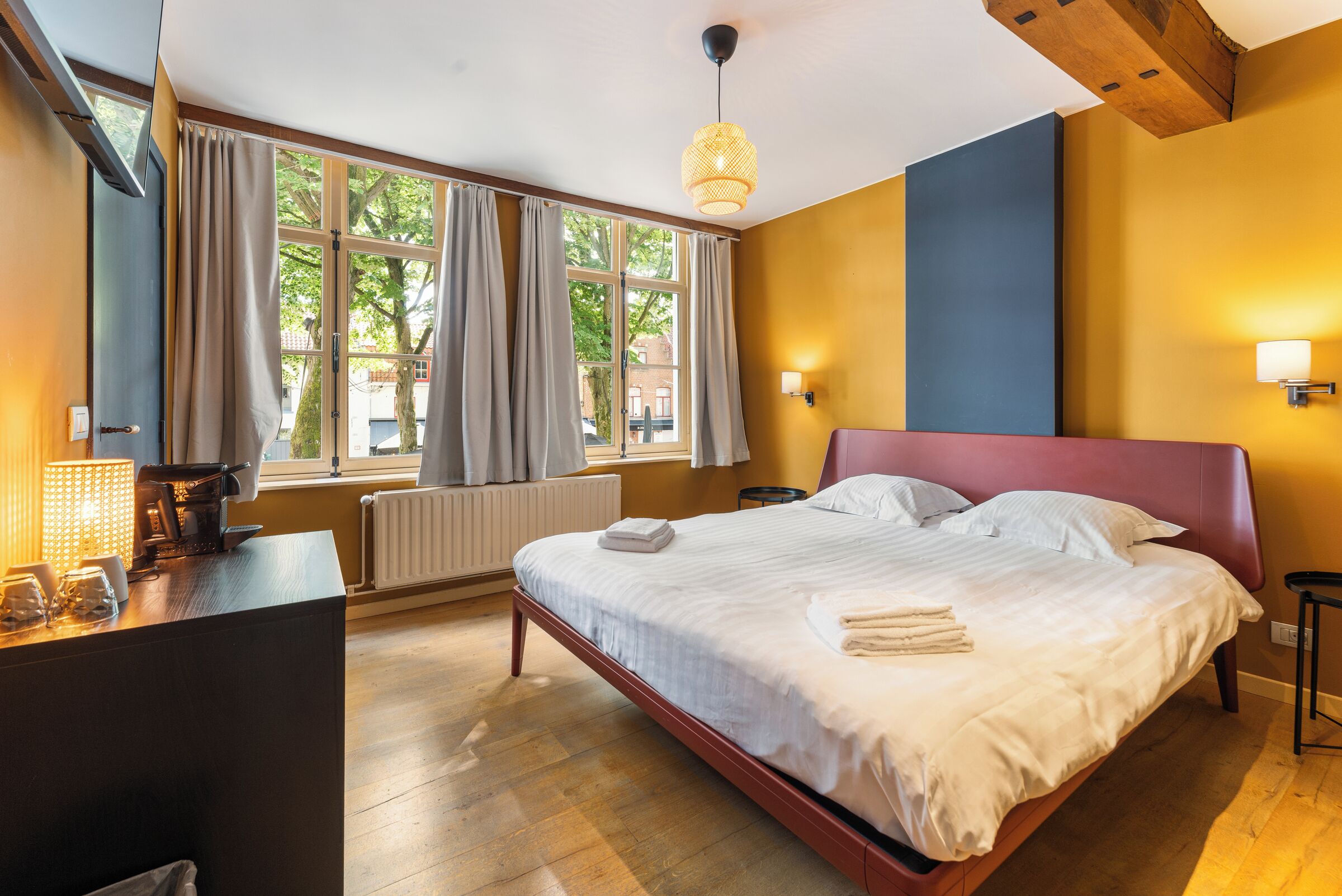 A hotel room with a bed, a desk, a window with curtains, a radiator, and a wall-mounted light fixture.