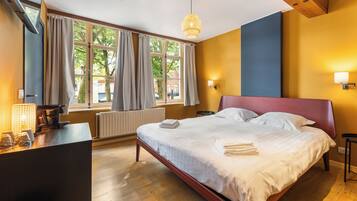 A hotel room with a bed, a desk, a window with curtains, a radiator, and a wall-mounted light fixture.