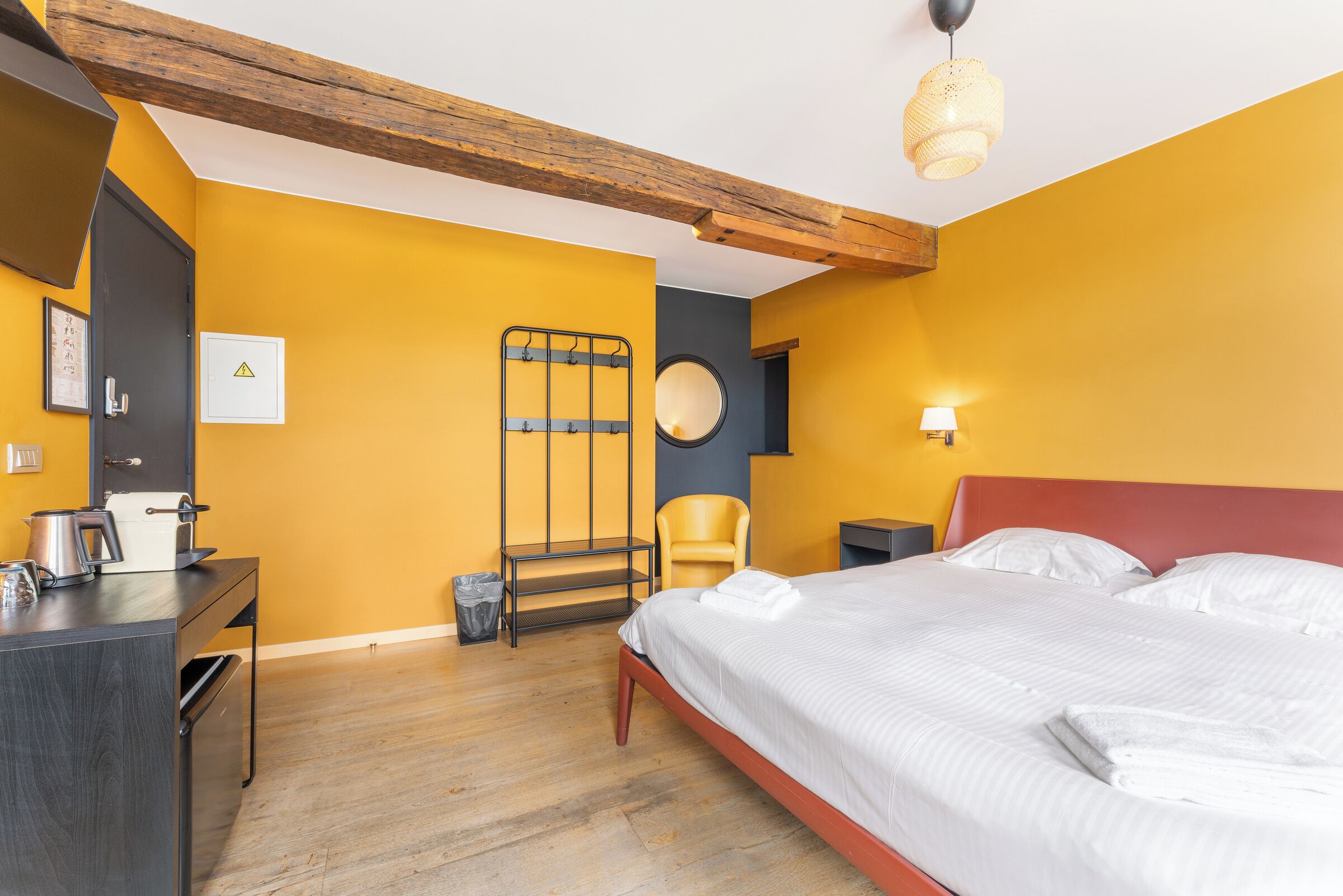 A hotel room with a bed, a desk, a chair, a small table, a trash bin, a wall-mounted shelf, and a wall-mounted round mirror.