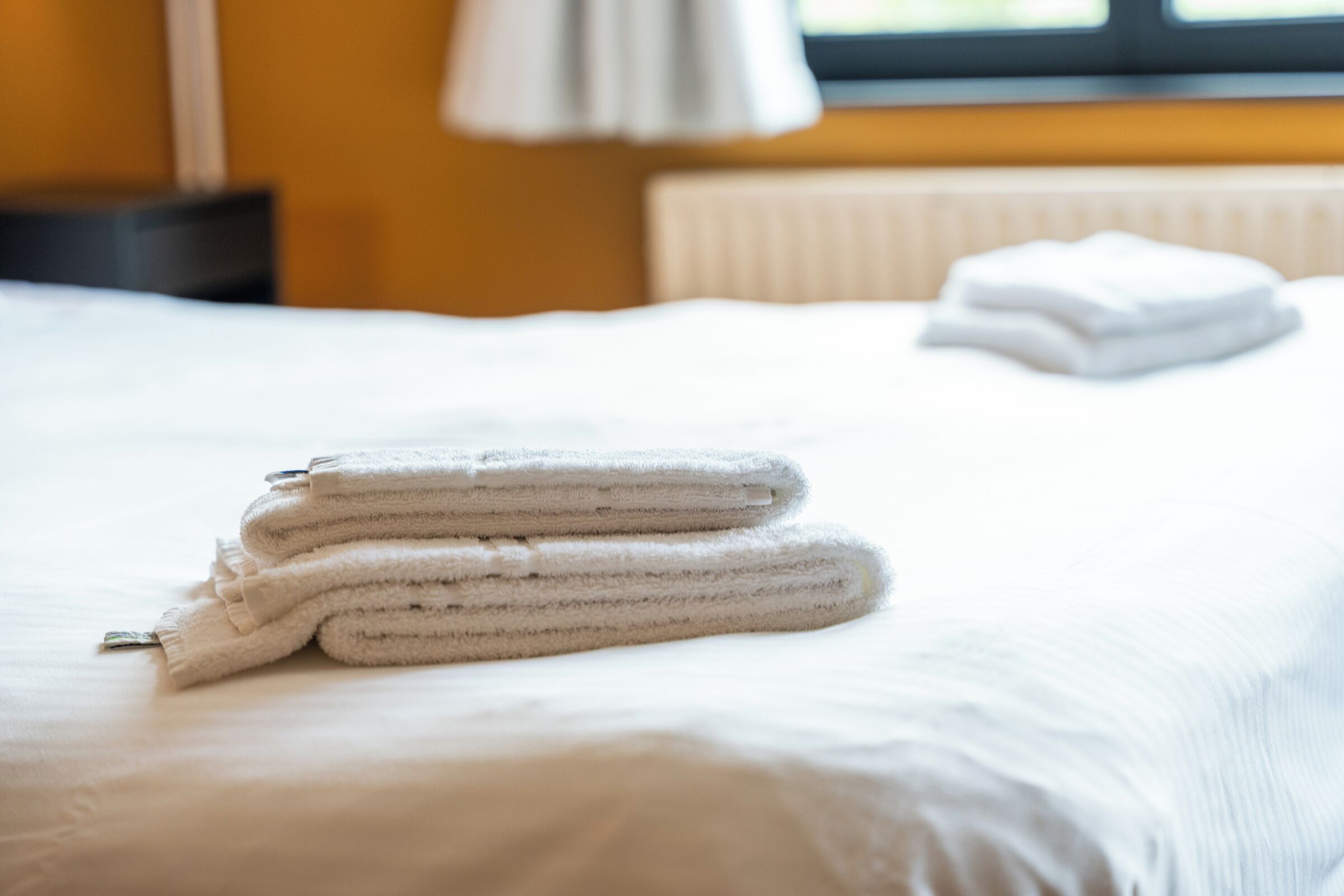 A neatly made bed with folded white towels and a window with blinds in the background.