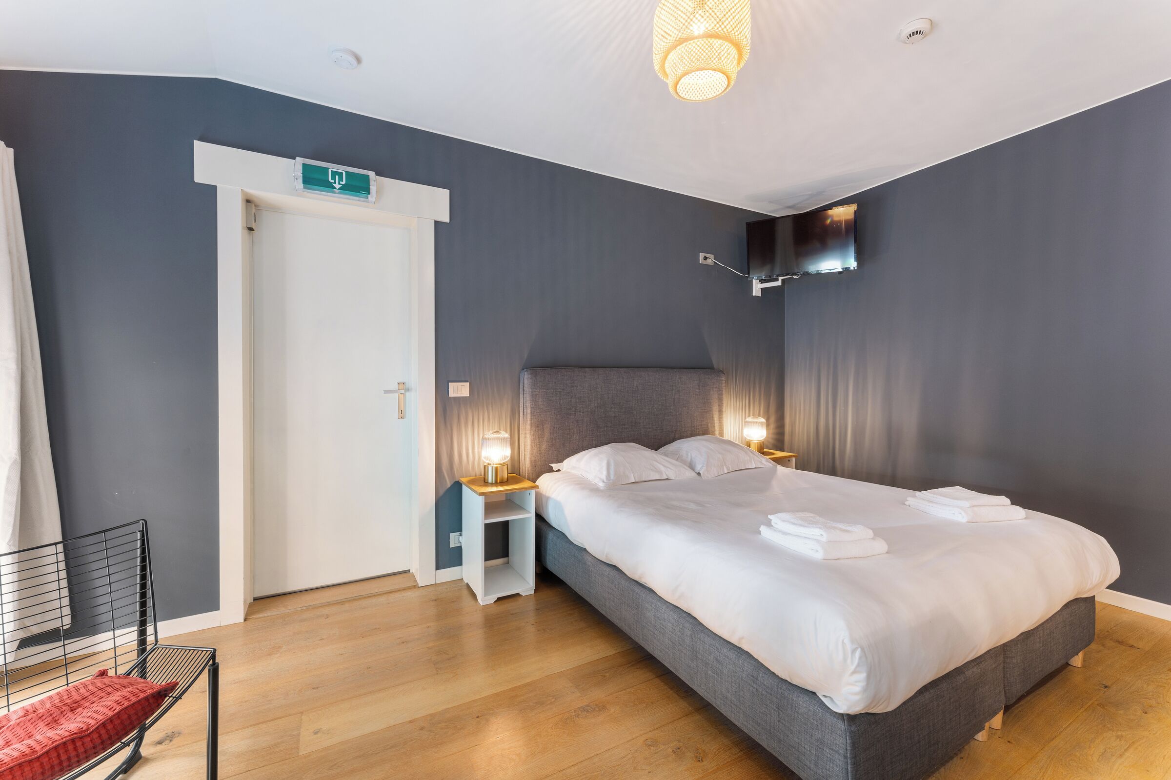 A modern hotel room with a large bed, bedside lamps, a flat-screen TV, and a door with an emergency exit sign.