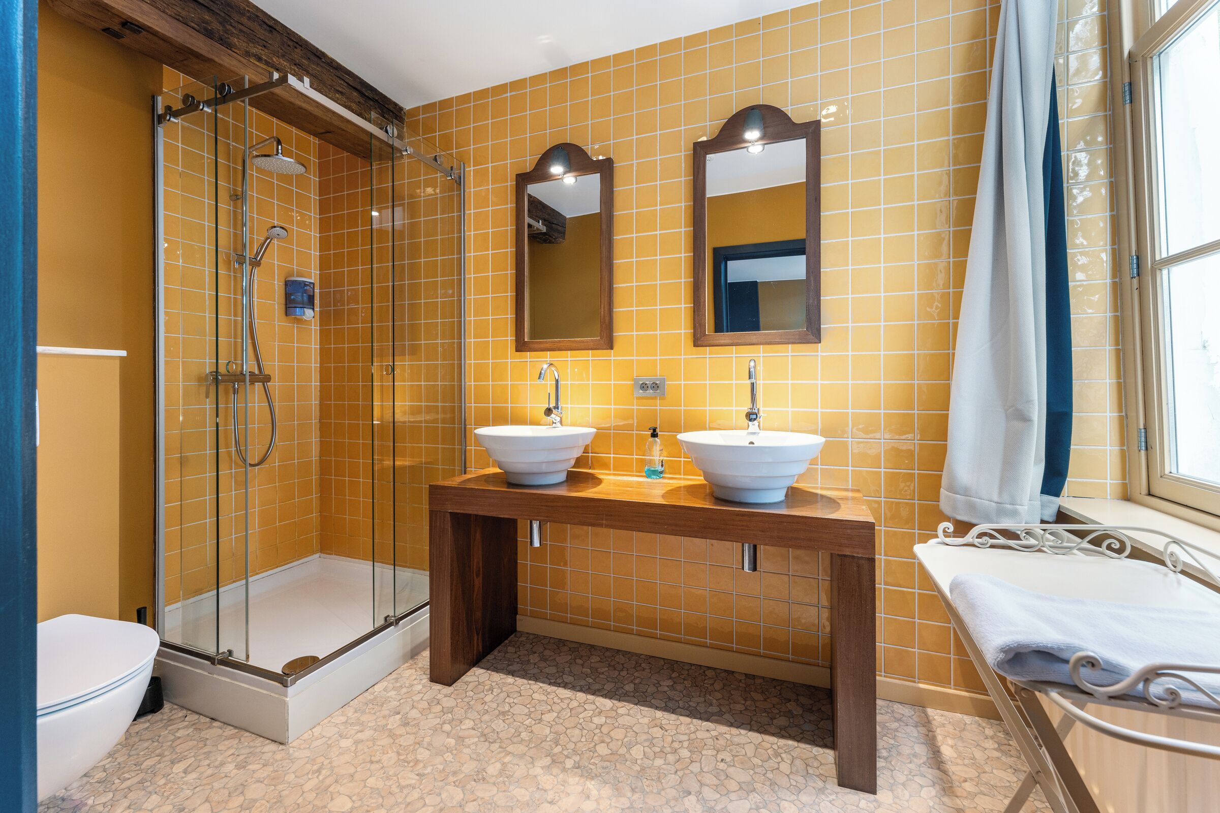 A bathroom with two sinks, a glass-enclosed shower, and a window with a curtain.