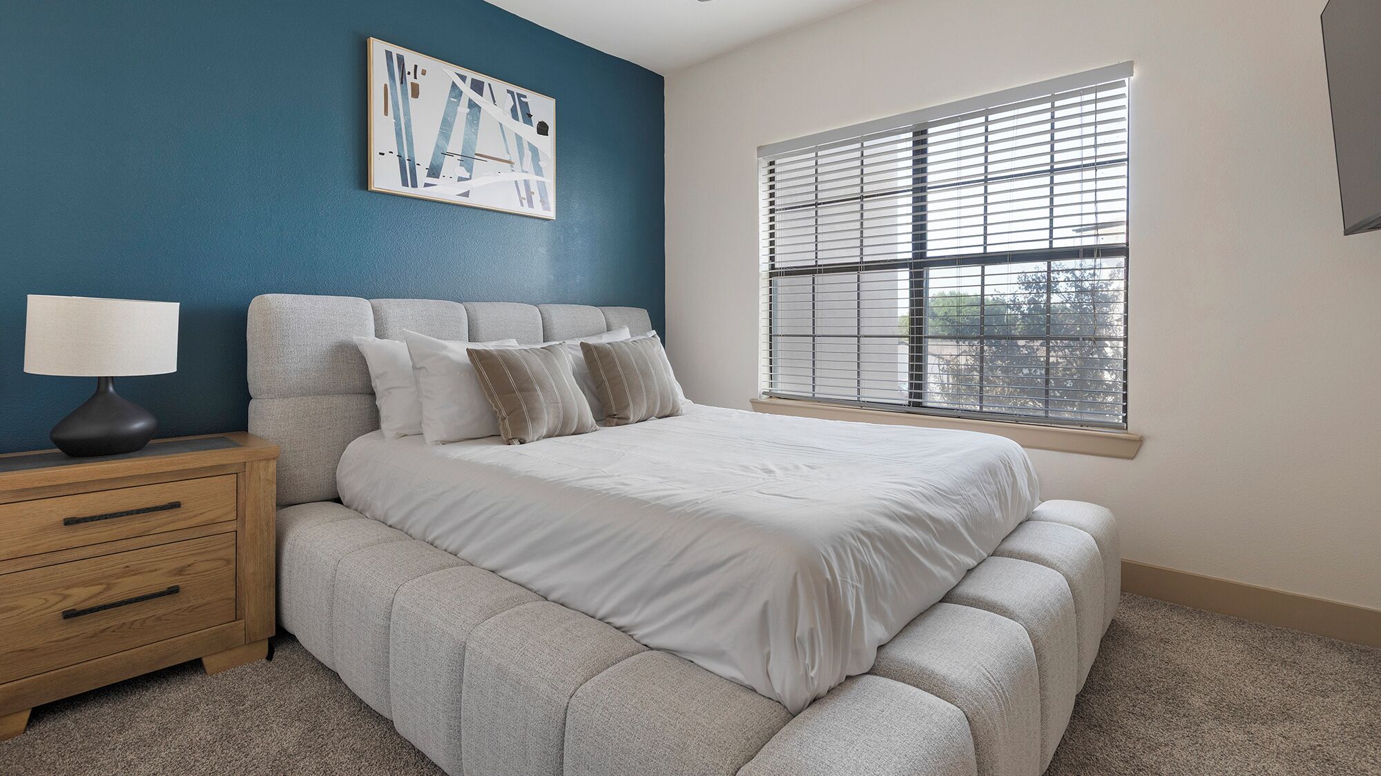 Un dormitorio con cama, mesita de noche, lámpara, ventana con persianas y una pared de acento azul.