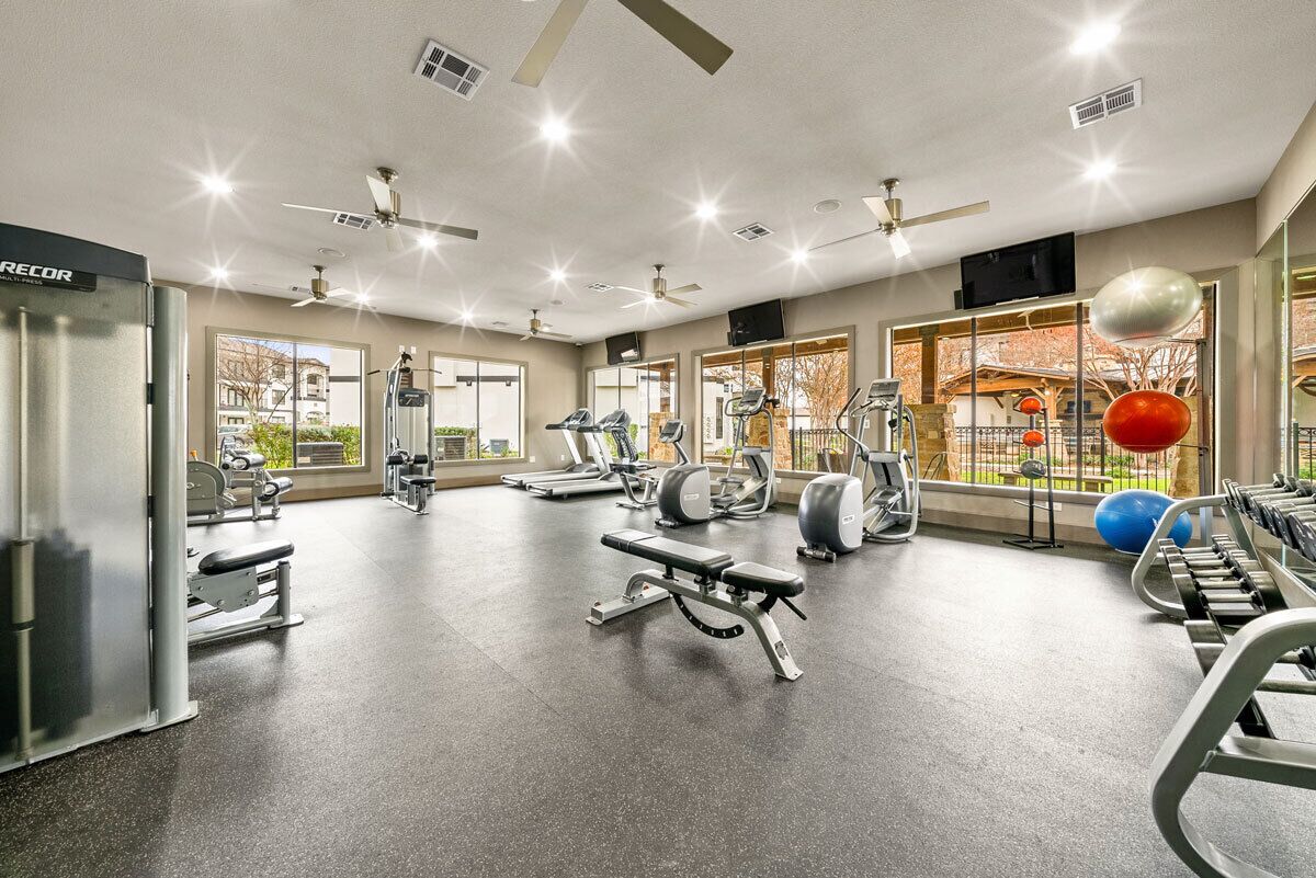 A modern gym with various exercise equipment, large windows, and ceiling fans.