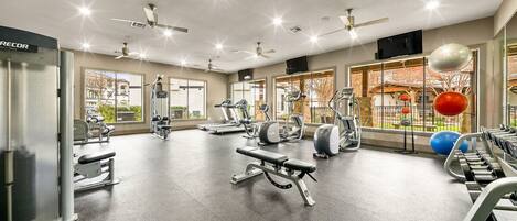 A modern gym with various exercise equipment, large windows, and ceiling fans.