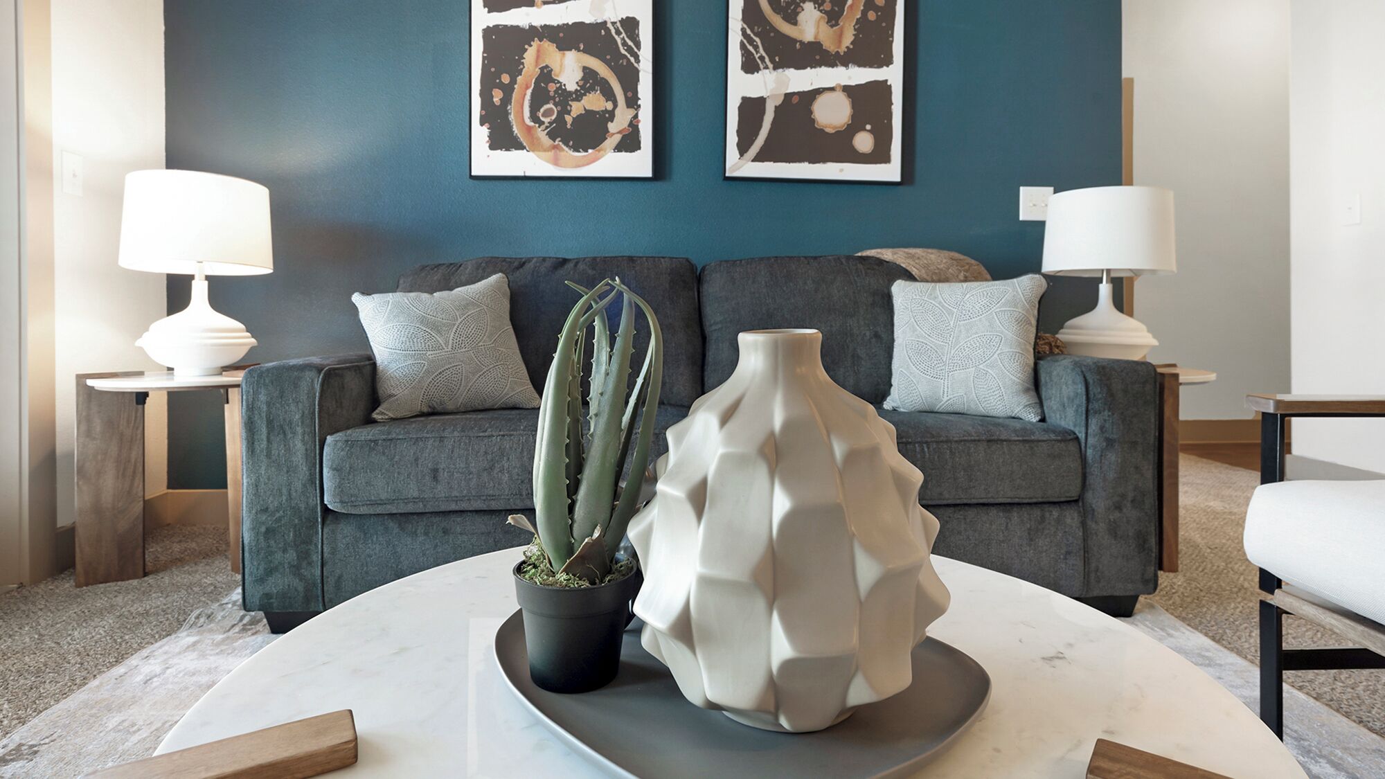 A modern living room with a grey sofa, two side tables with lamps, a marble coffee table, a potted cactus, and a unique white vase.