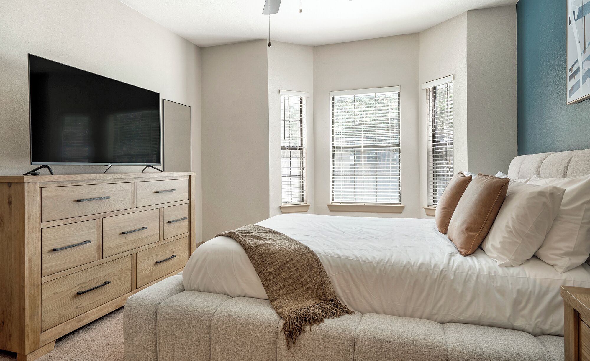 A bedroom with a bed, a TV on a wooden dresser, and windows with blinds.