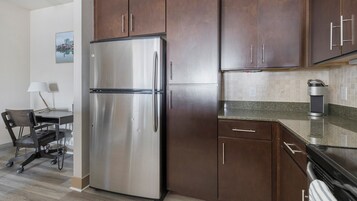 A compact kitchen with a stainless steel refrigerator, dark wooden cabinets, a granite countertop, and a coffee maker.