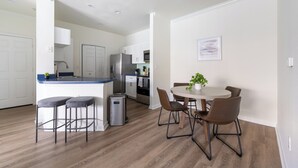Een moderne keuken met witte kasten, een roestvrijstalen koelkast en een blauw aanrecht. Er is een zithoek met een ronde tafel en vier stoelen, en een kleine potplant op de tafel.