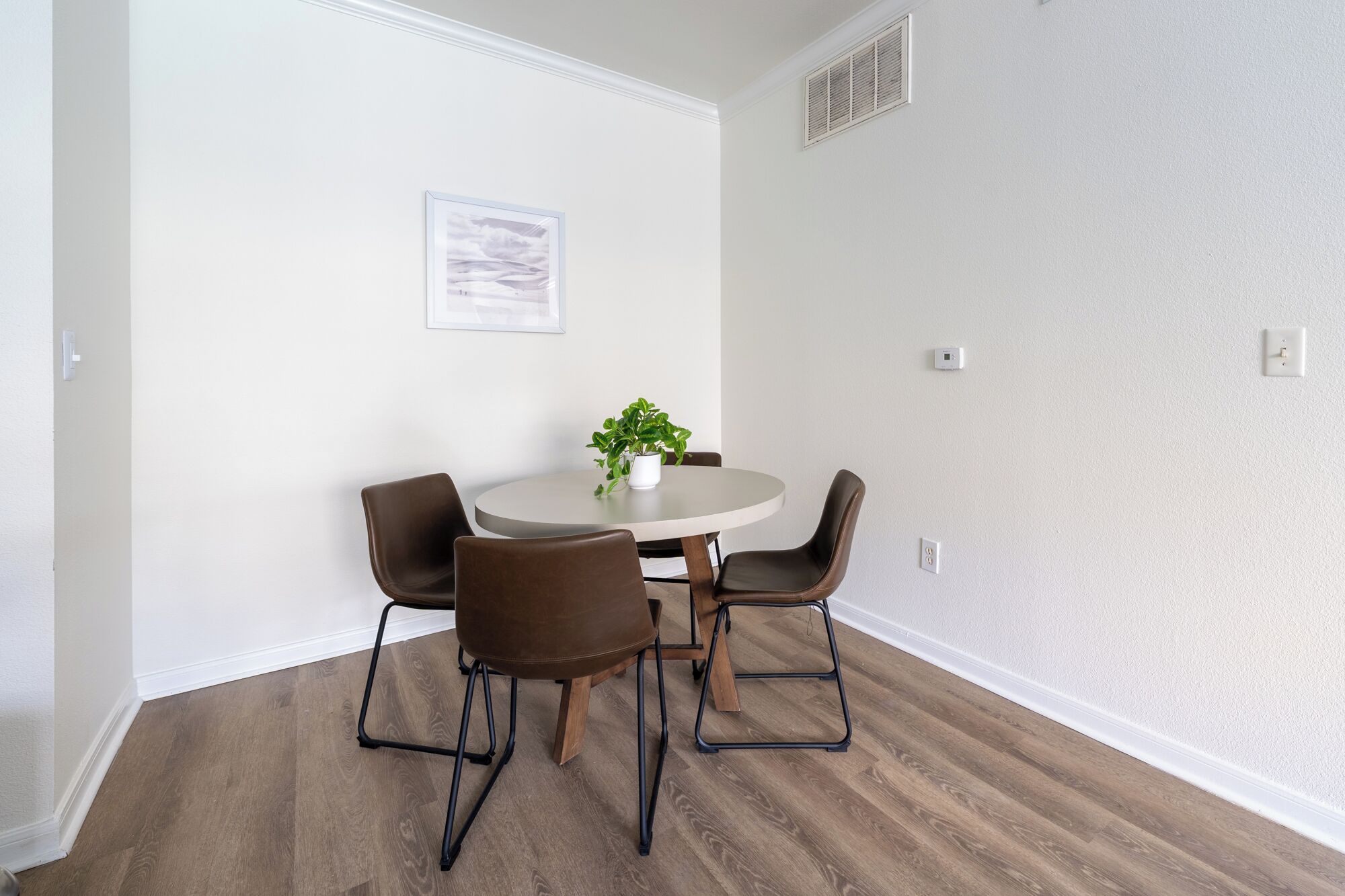 A dining area with a round table, four chairs, a potted plant, and a framed picture on the wall.
