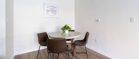 Uma sala de jantar com uma mesa redonda, quatro cadeiras, um vaso de planta e um quadro na parede.
