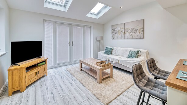 A modern living room with a flat-screen TV, wooden entertainment unit, white sofa, and a dining area with bar stools. - One Bed in Guilden Sutton by Guestz (Guilden Sutton)