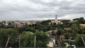 A view of a cityscape with a prominent church featuring a tall bell tower, surrounded by lush greenery and residential buildings. - Hello Roma 2 (Rome)