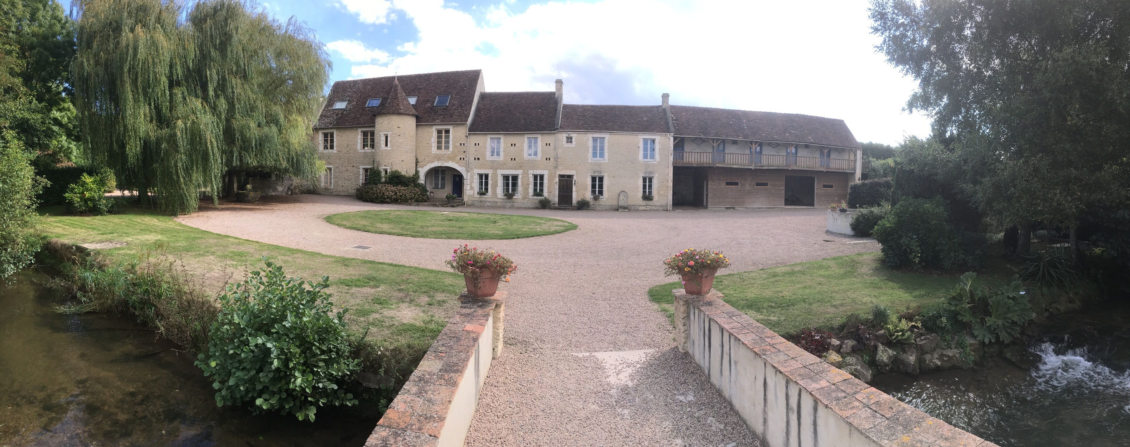 A large stone building with a courtyard, a small bridge over a stream, and a weeping willow tree.
