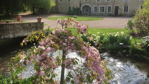 A courtyard with a stone bridge, a pond with pink flowers, and a large building with multiple windows and a sloped roof. - Coeur de Combray, sleeps 44 with pool & function space set in beautiful gardens (Ernes)