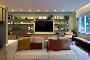 A living room with a sofa, TV, shelves with decorative items, and a window. - Ipanema | Duplex no melhor local do bairro, 4 quartos, 5 min da Praia - por Casapē Rio (Rio de Janeiro)