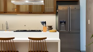 A modern kitchen with wooden cabinets, a stainless steel refrigerator, a white countertop, and two wooden bar stools. - Ipanema | Duplex no melhor local do bairro, 4 quartos, 5 min da Praia - por Casapē Rio (Rio de Janeiro)
