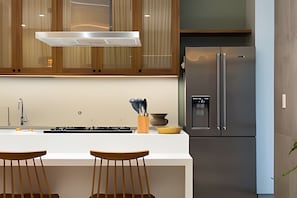Une cuisine moderne avec des meubles en bois, un réfrigérateur en acier inoxydable, un plan de travail blanc et deux tabourets de bar en bois.