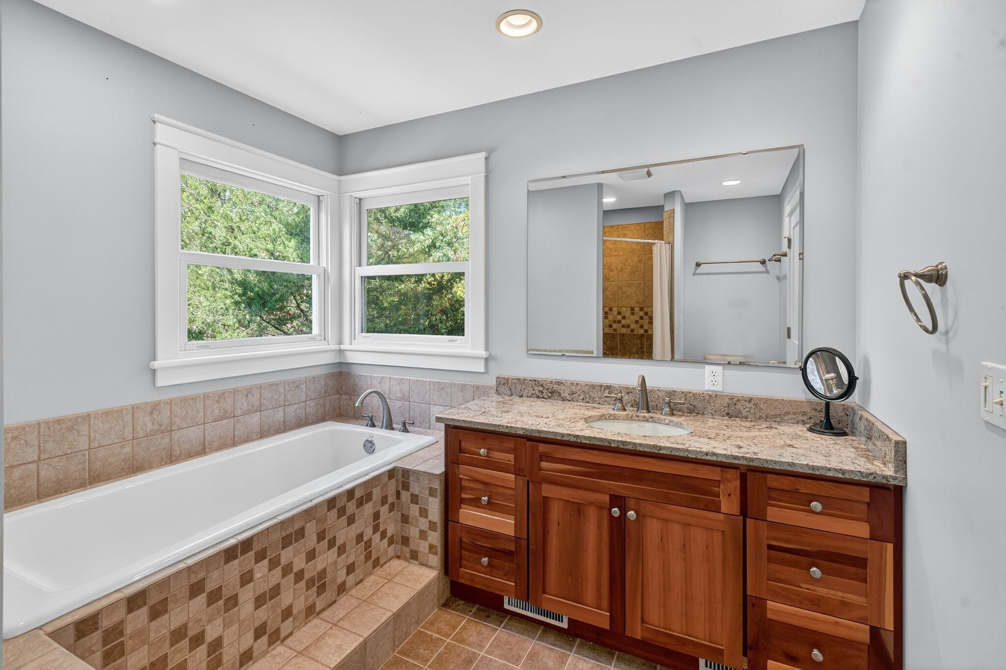 A modern bathroom with a freestanding bathtub, wooden vanity with drawers, a large mirror, and a window with a view of greenery.