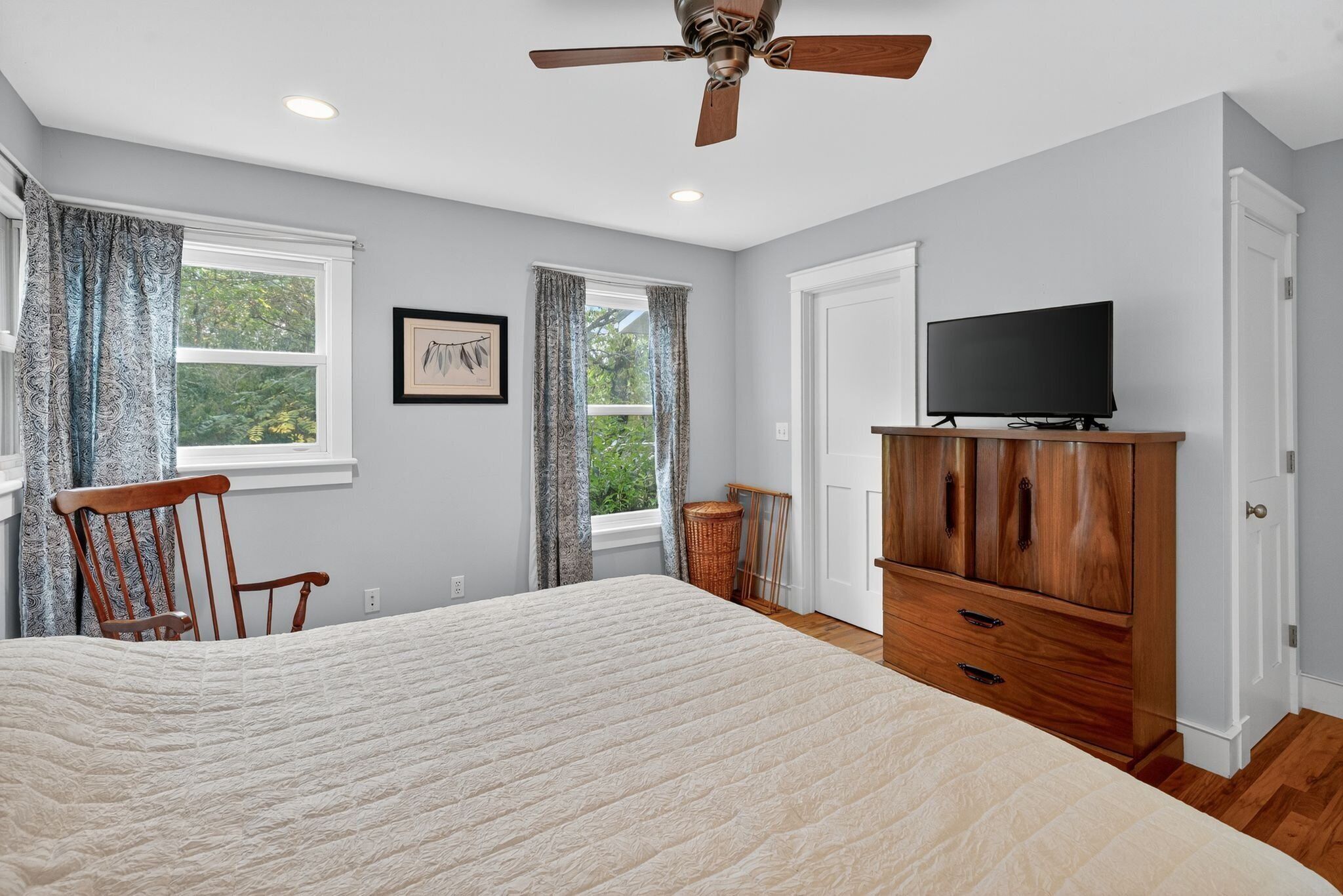 A bedroom with a large bed, a wooden wardrobe, a television, and two windows with curtains.