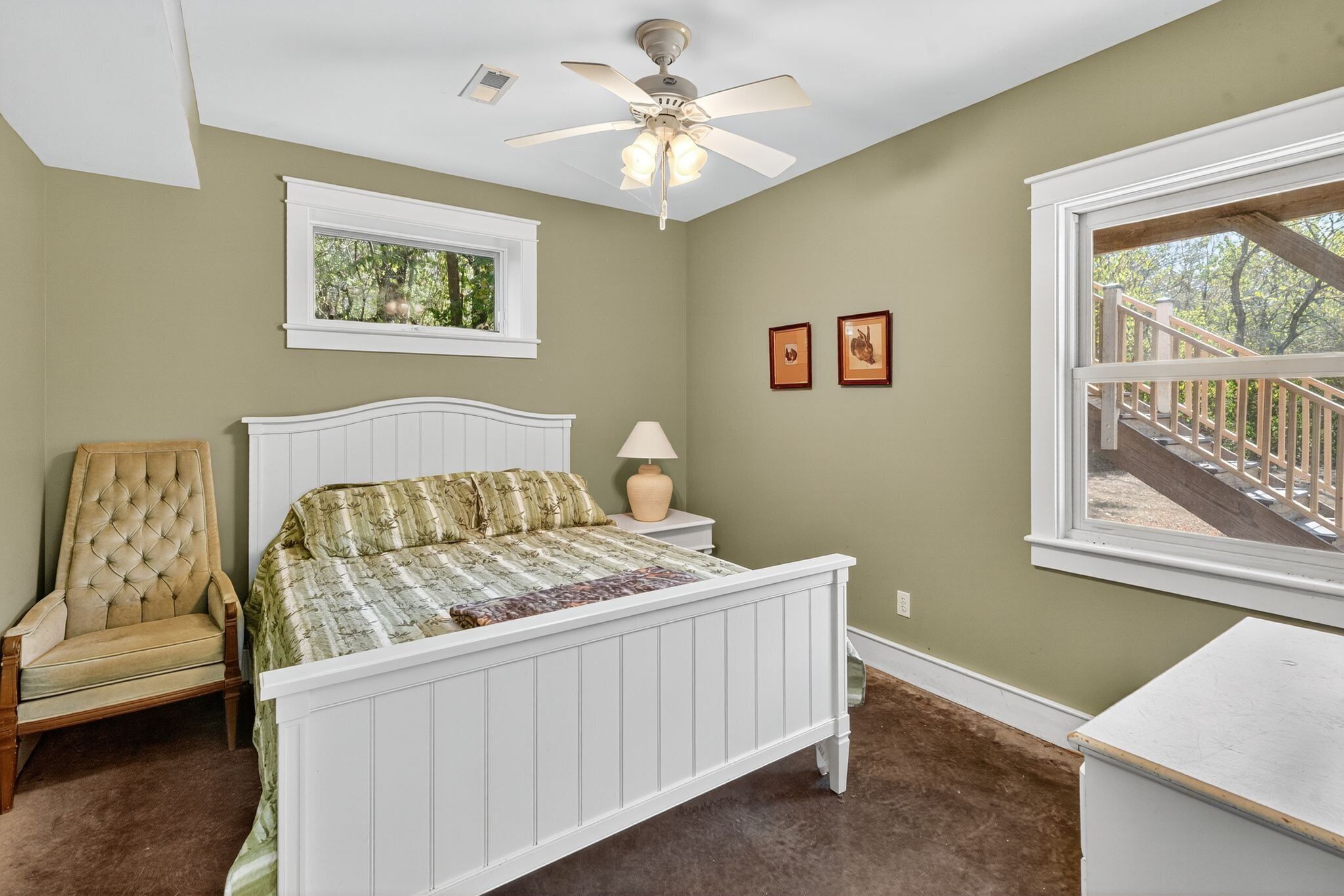 A bedroom with a bed, a chair, a nightstand, a lamp, a ceiling fan, a window with a view of a staircase, and two framed pictures on the wall.