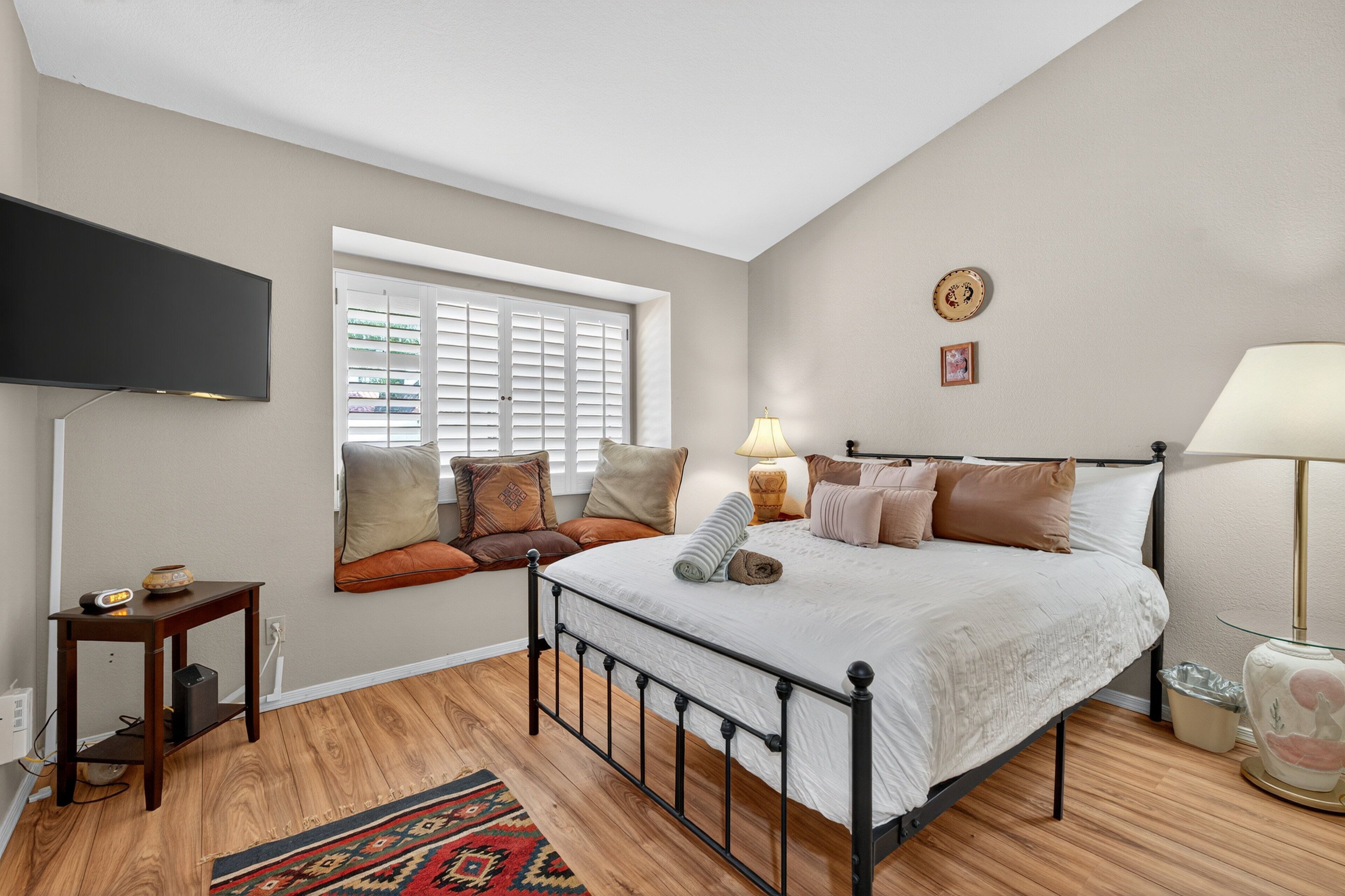 A bedroom with a bed, a window with blinds, a TV mounted on the wall, a bedside table, and a lamp.