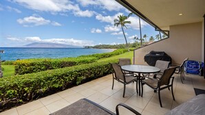 A patio with a round table and chairs, a grill, and a view of the ocean and mountains. - Makena Surf, #f-111 2 Bedroom Condo (Kihei)