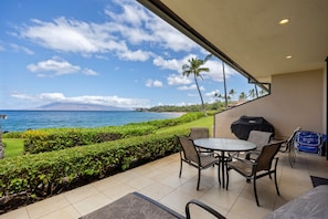 A patio with a round table and chairs, a grill, and a view of the ocean and mountains. - Makena Surf, #f-111 2 Bedroom Condo (Kihei)