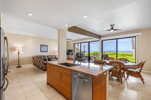 A modern kitchen with a large island, a dining area with wicker chairs, and a view of the ocean. - Makena Surf, #f-109 2 Bedroom Condo (Kihei)
