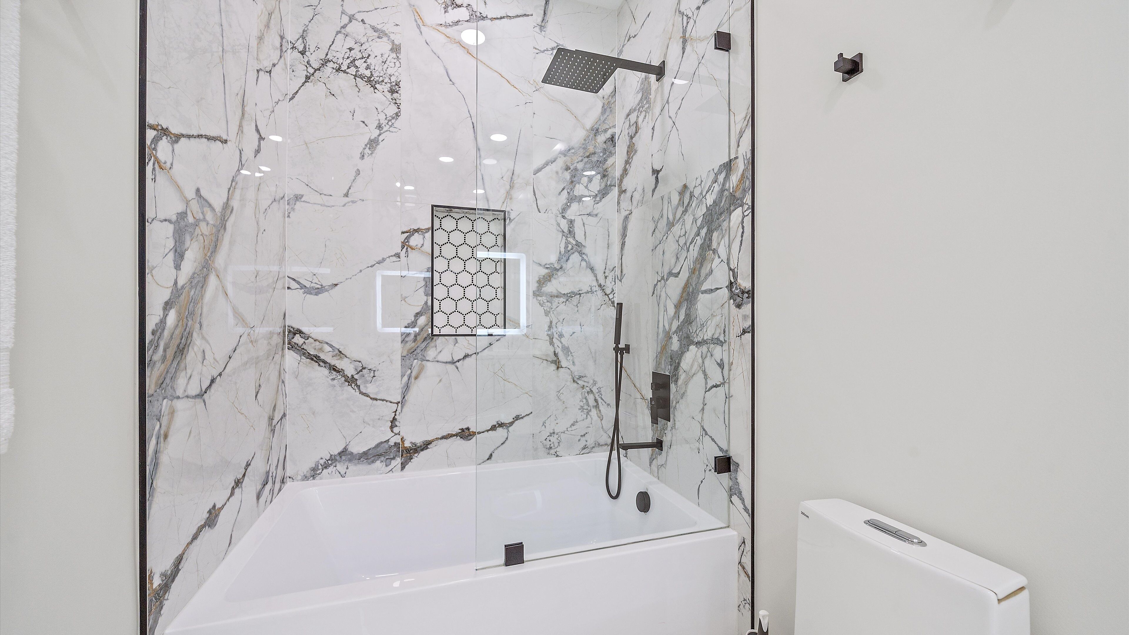 A modern bathroom with a marble wall, a glass-enclosed shower, and a white bathtub.