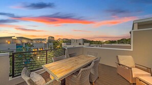 Eine Dachterrasse mit Holztisch und Korbstühlen mit Blick auf eine Stadtlandschaft bei Sonnenuntergang.