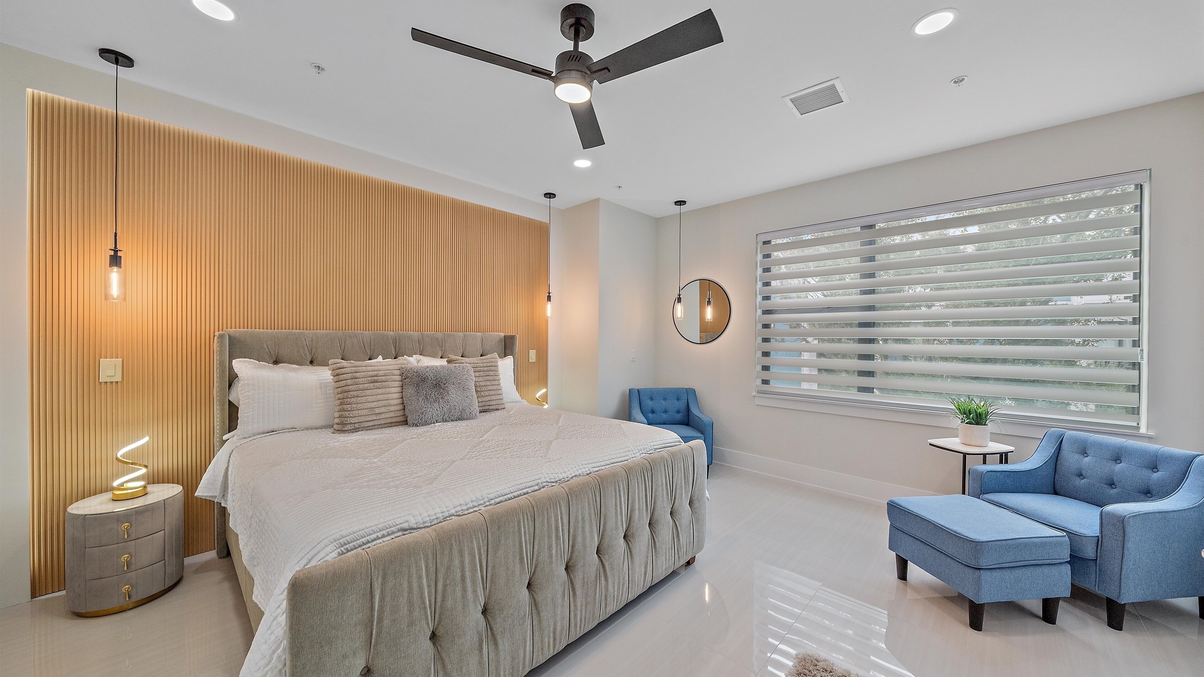 A modern bedroom with a large bed, a ceiling fan, a window with blinds, and a blue armchair.