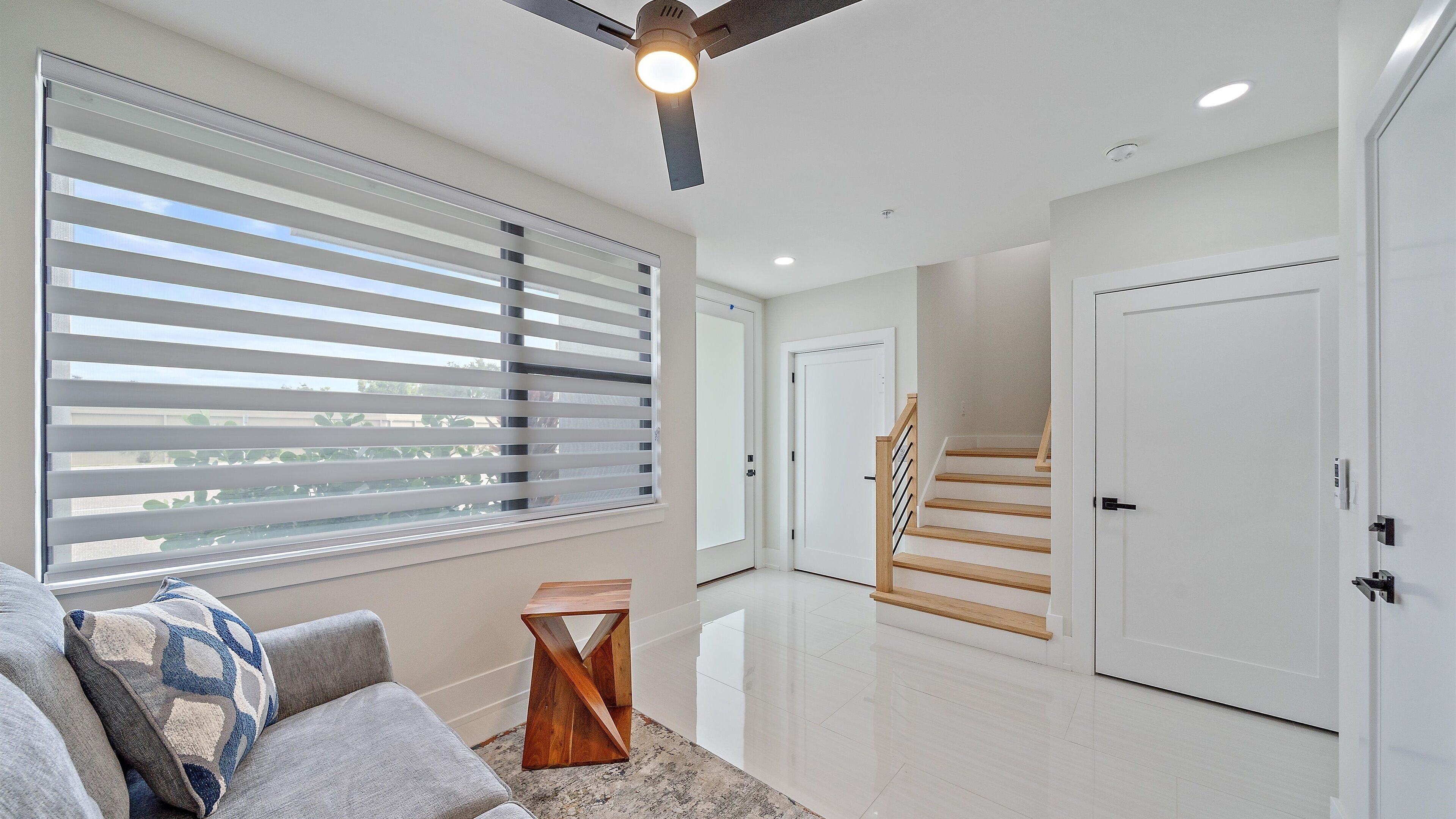 A modern living room with a large window, a ceiling fan, and a staircase leading to another level.