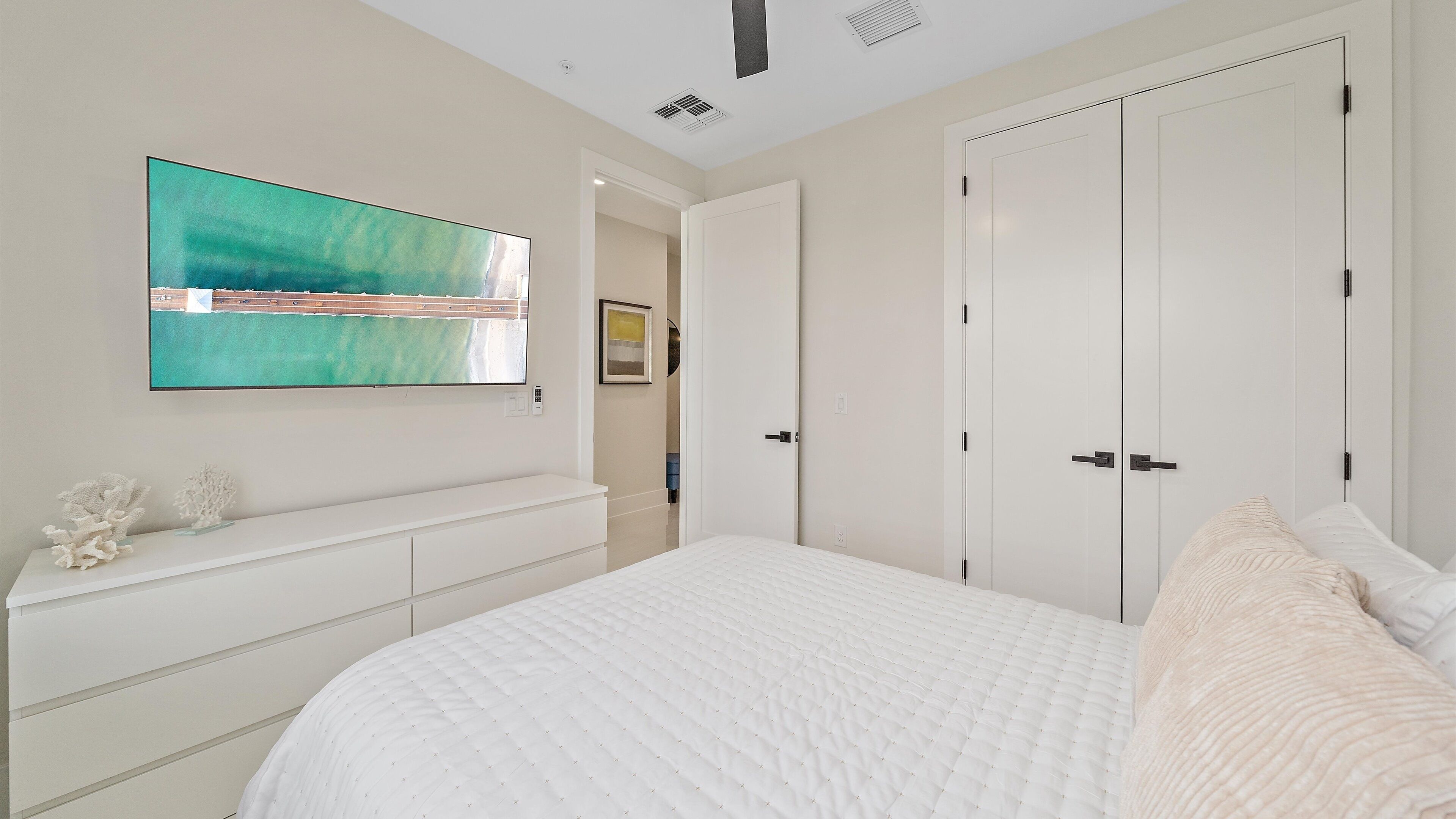 A bedroom with a large bed, a white dresser, a flat-screen TV mounted on the wall, and a door leading to another room.