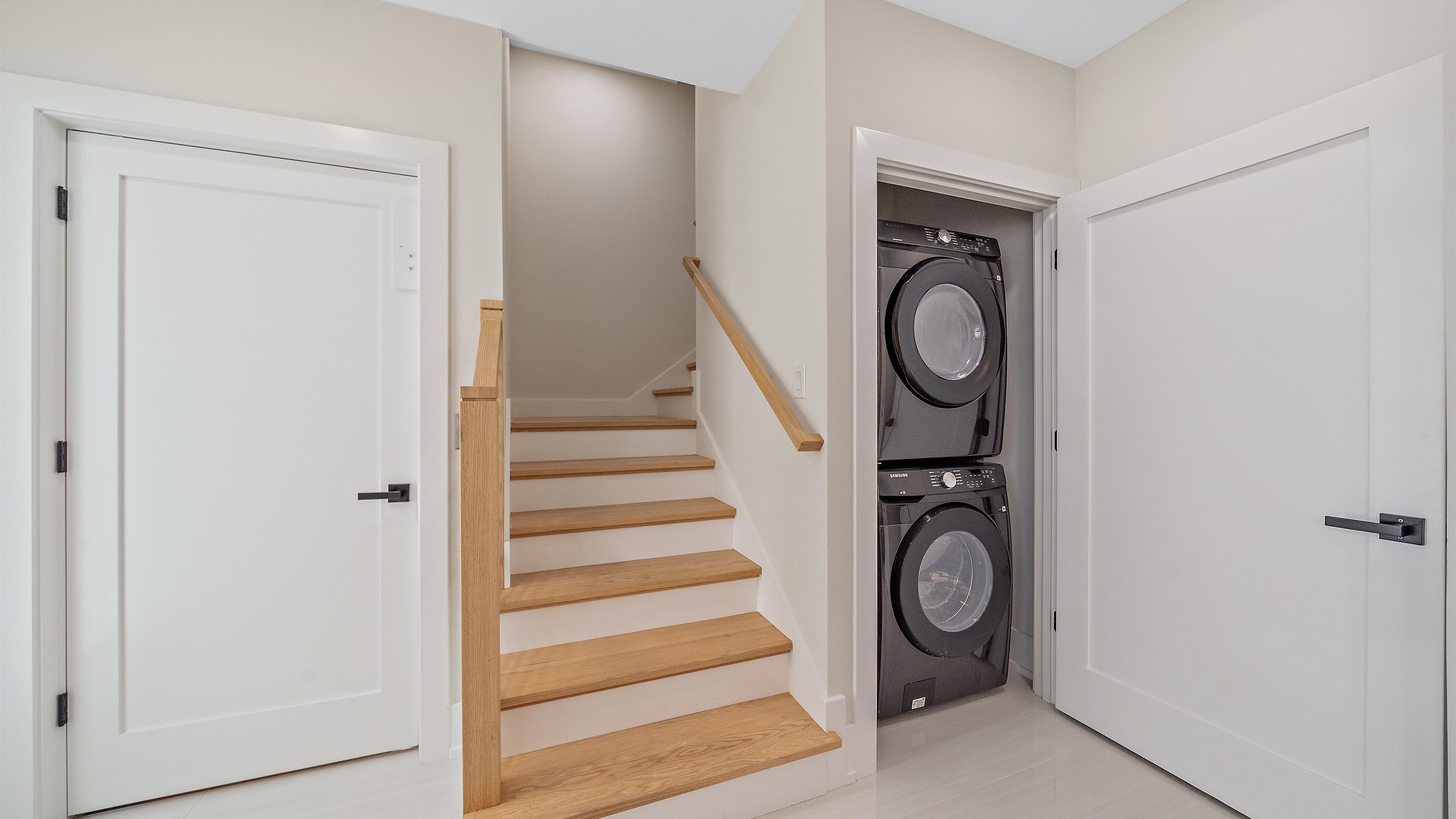 A clean, modern hallway with white walls and wooden stairs leading to a laundry area with stacked washing machines.