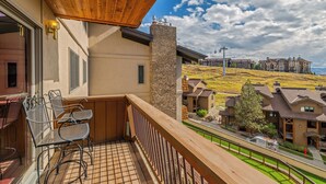 A balcony with a wooden ceiling, metal chairs, and a view of a residential area with houses and a grassy hill. - Storm Meadows I -1-Bedroom + Studio (Retractable Privacy Wall), 2-Bath (Steamboat Springs)