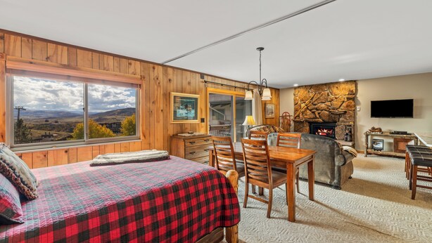A cozy room with a bed, wooden walls, a fireplace, a dining table, and a TV. - Storm Meadows I -1-Bedroom + Studio (Retractable Privacy Wall), 2-Bath (Steamboat Springs)