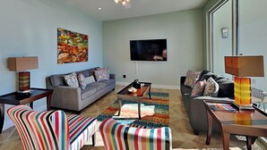A modern living room with a grey sofa, striped chairs, a coffee table, and a TV on the wall. - Diamond Beach #214 | Ocean Views, Resort Pool (Galveston)