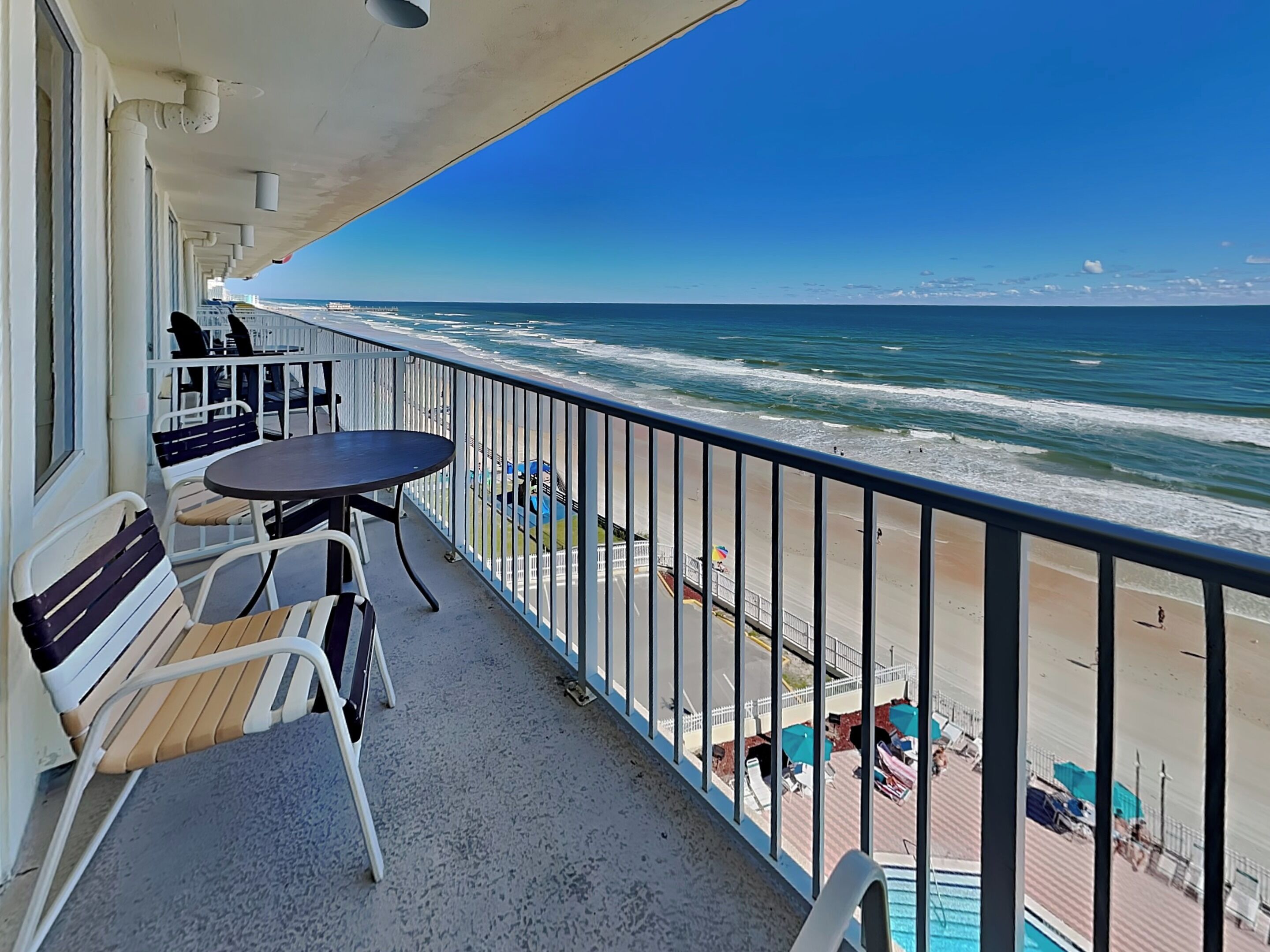 A balcony with a table and chairs, overlooking a beach with umbrellas and a pool.