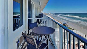 Un balcon avec une table et des chaises, donnant sur une plage où l’on aperçoit des vagues et, au loin, une jetée.