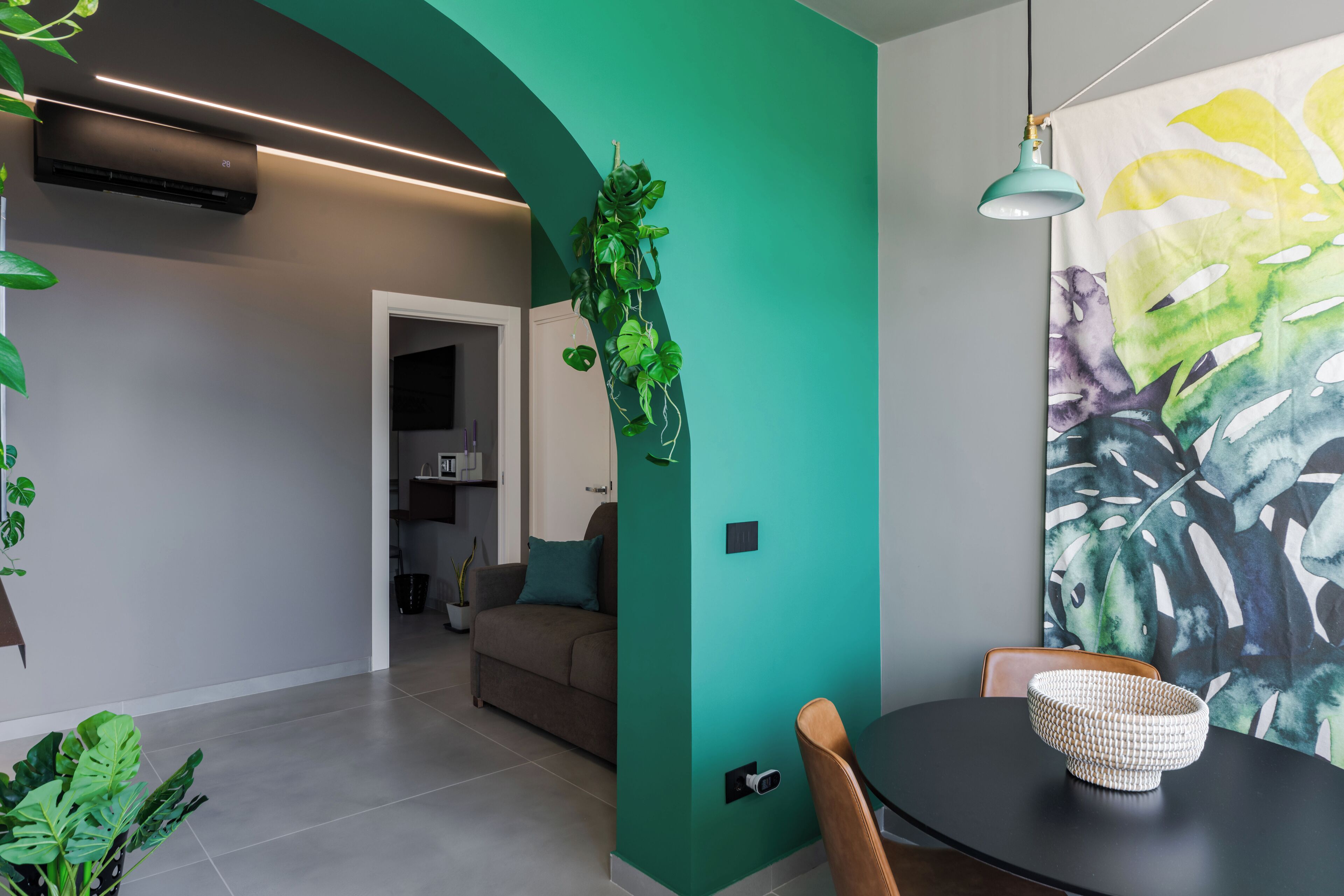 A modern living room with a teal accent wall, a dining area with a black table and a patterned bowl, and a hanging plant.