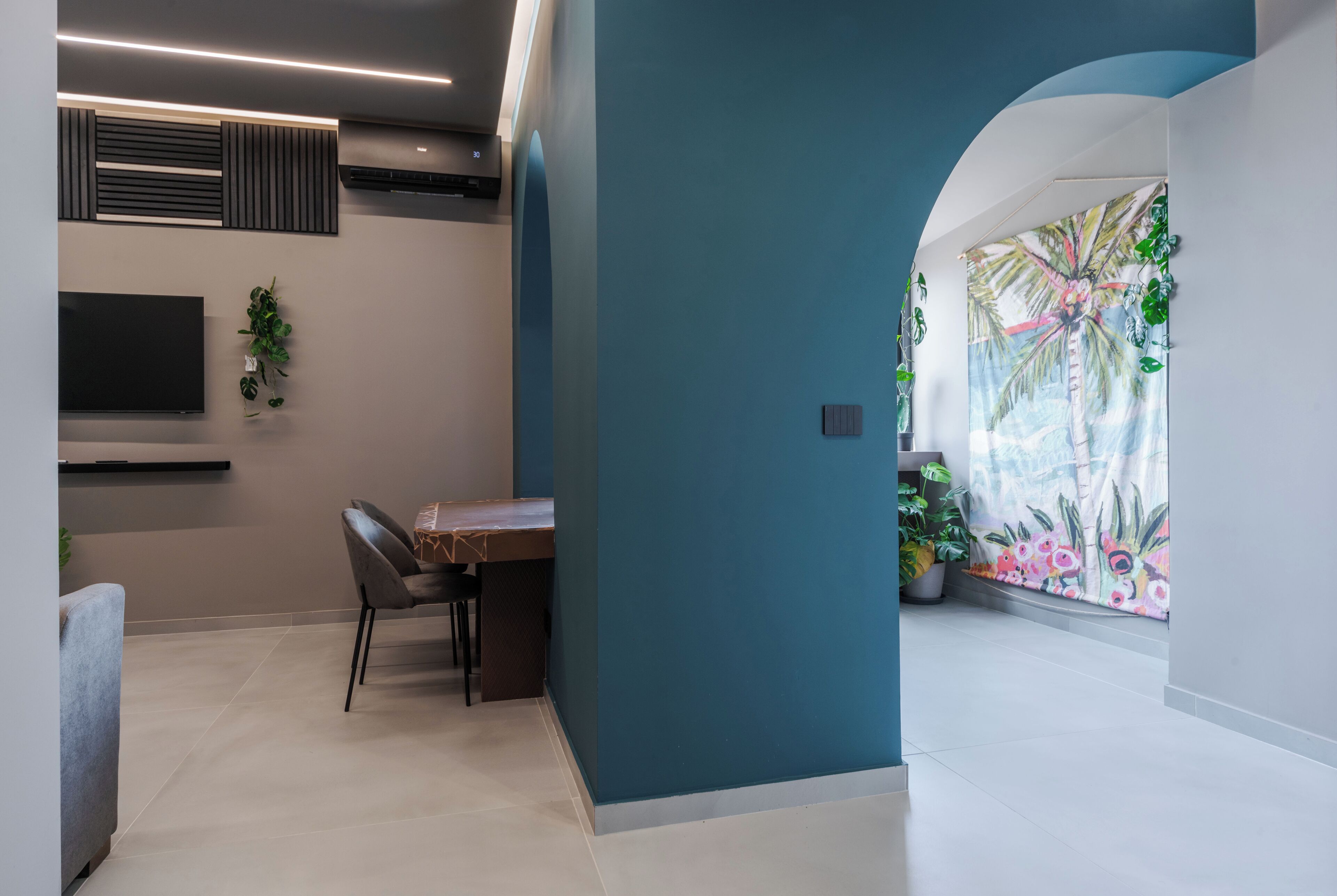A modern living room with a flat-screen TV, a dining table with chairs, and a large window with tropical plants.