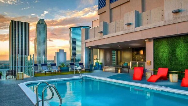 A rooftop pool with lounge chairs and a bar area, surrounded by modern buildings and a sunset view.