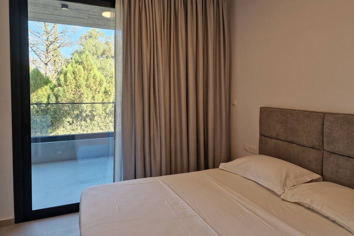 A bedroom with a large window, a balcony, and a bed with white bedding.