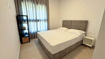 A modern bedroom with a large bed, a nightstand, a bookshelf, and a window with curtains.