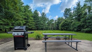 A barbecue grill and picnic table set on a concrete surface with trees in the background. - The Pines at New Albany | Sleeps 8 | Acre Lot (New Albany)