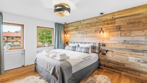 A modern bedroom with a wooden wall panel, a large bed with white linens, two pendant lights, and a view of a building through a window. - Suite Gipfelstürmer | Pool & sauna and mountain view (Reit im Winkl)